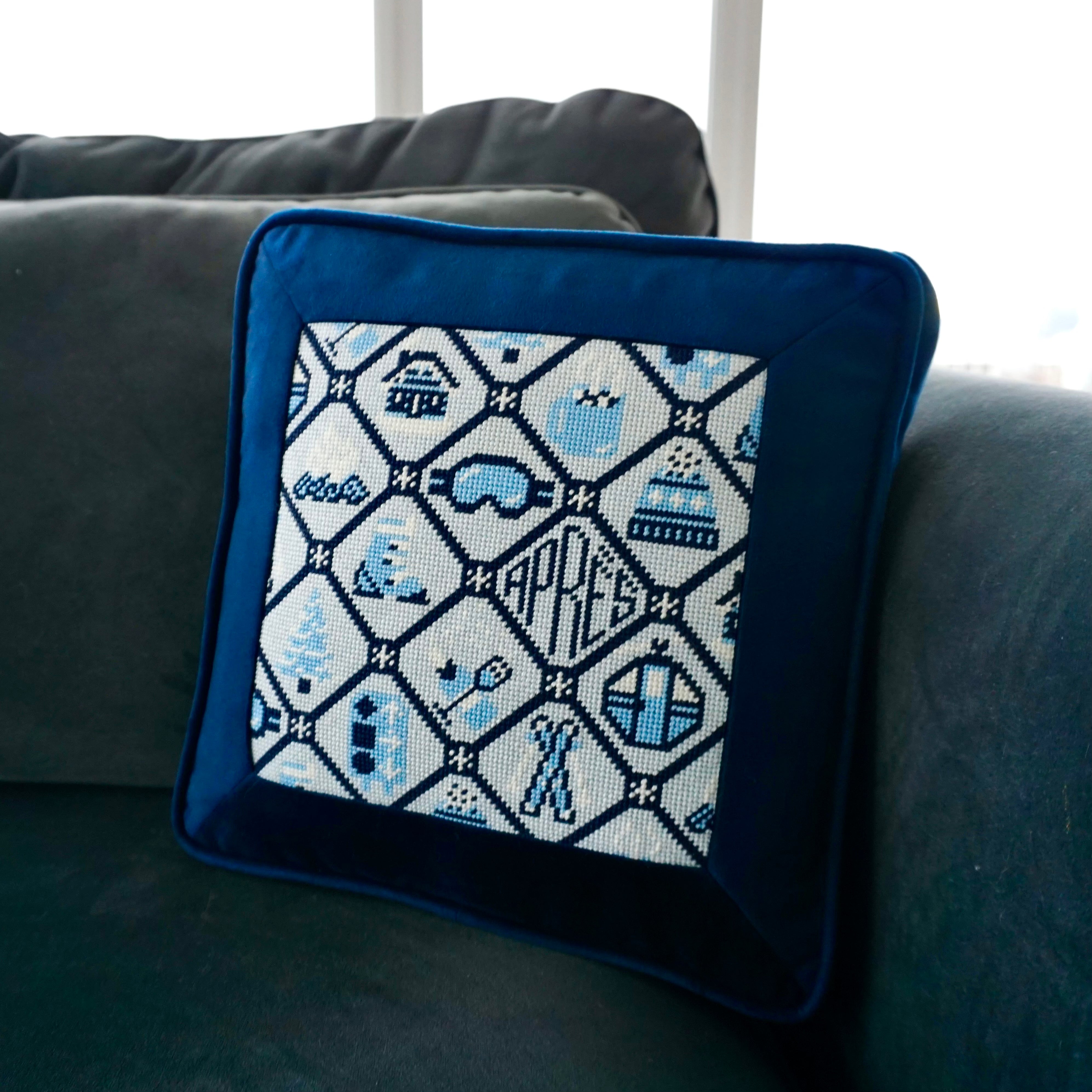 Après ski needlepoint canvas featuring apres ski themed icons including skis, campfire, cocoa, gondola, champagne, and more finished into a pillow with blue velvet